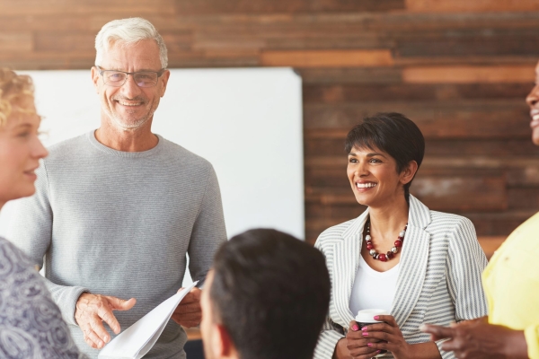 One white haired man wearing a light grey jumper talks to three woman (blond white woman in grey top, Asian woman with dark hair in grey jacket and black woman in a yello top) in an office setting with the back of another mans head visible at the bot